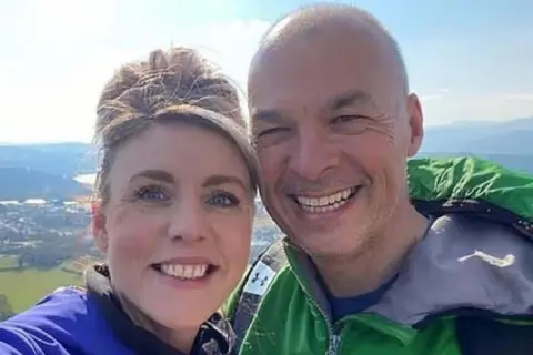 Rebecca Hutt (left) and Det Supt Steven Williams (right) are photographed together from the shoulders up. They are both smiling and looking at the camera. They are outdoors on a sunny day at the top of a high view point. 