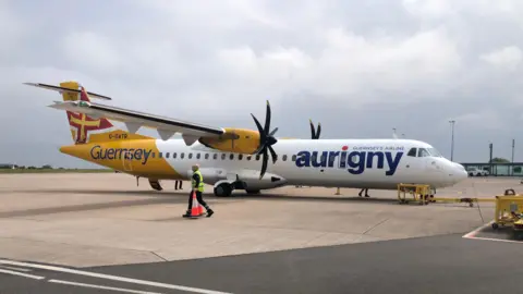 BBC An Aurigny ATR plane on the runway at Guernsey Airport. It has a white and yellow livery and propellers.