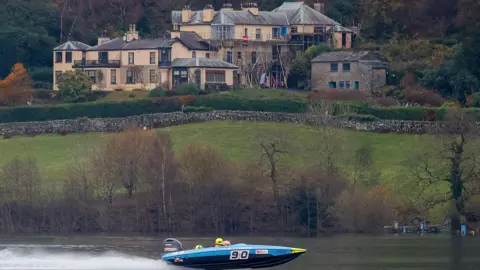 John Heald Photography Light blue boat with the number 90 written on the side. There are two people wearing helmets inside the boat and it is moving at speed through the water. There is water spray on the left-hand side of the photo. There is a large building covered in scaffolding in the background and a number of trees and bushes.