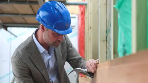 The Prince of Wales poises a hammer over a nail on a building site, he wears a blue hard hat 