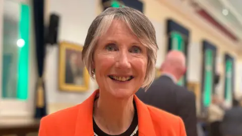 BBC Fiona is wearing a bright orange blazer and a black top stands indoors at what appears to be a formal or social event. She has a name badge pinned to the blazer and a pearl-style necklace. The background shows a large hall with wooden flooring, blue chairs arranged in rows, tall windows with dark curtains, framed pictures on the walls, and several other attendees minglin
