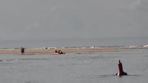 Two people on a sandbank in the middle distance as a lifeboat approaches. The is water between two separate areas of sandbank. A red buoy is floating in the foreground on the right.