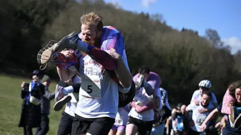 A group of people carrying others on their shoulders. Some are giving the person they are carrying a piggy back, others are carrying with the person's feet in front of them, and them hanging down their back.