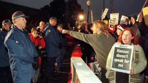 Getty Images Protesters hold signs reading "Hanson doesn't speak for me" behind a white barricade, behind which are police officers.