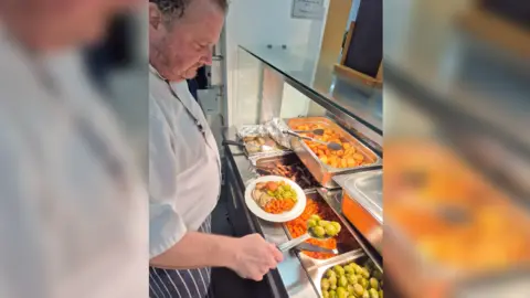 inHope A man in a striped apron and white shirt serves a roast dinner from a serving hatch. 