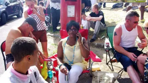 KAREN STODDART A woman in a yellow sleeveless top and white leggings sitting down on a fold-up chair holding a mobile phone to her ear surrounded by other people.