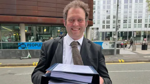 Dr Mick Salt, a man with light brown hair, is stood by the side of a road, wearing a grey suit jacket and white shirt with a brown tie. He is holding a number of large lever arch files full of papers.