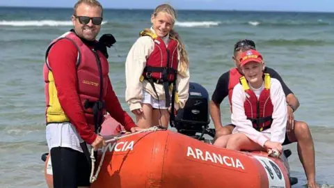 Two girls are wearing life jackets and are aboard a blow-up boat. The jackets are red. Annie is wearing a red hat. There are two adults next to them keeping the boat steady.