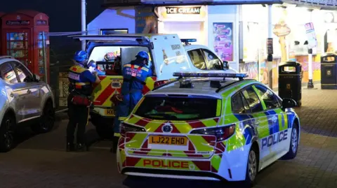 Eddie Mitchell Emergency responders wearing helmets stand beside an open search and rescue vehicle on Brighton seafront at night, with a marked police car in the foreground and a brightly lit ice cream kiosk and red telephone boxes behind them.