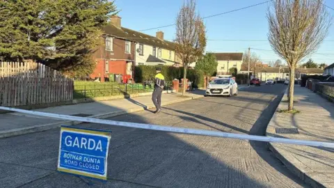 A residential street with houses and trees on either side of the road. Across the road is blue police tape and a sign in the middle of the road reads: "Gara, Bóthar Dúnta, Road Closed". A guard with his back to the camera is walking across the road and a garda car is parked on the street.