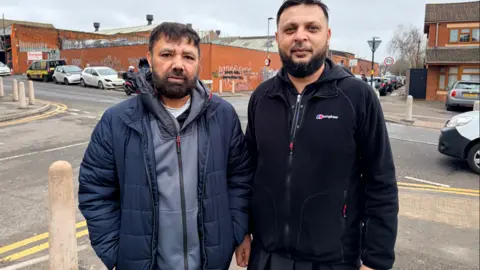 Two men stand on the corner of a residential street. One is wearing a grey hoodie and blue puffer jacket, while the other has a dark fleece top on. There is a crossroads behind them, showing cars parked on a pavement. 