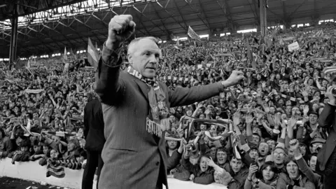 PA Media Black and white image shows Bill Shankly, in a suit and Liverpool scarf, turning towards the Kop end of Anfield to receive an ovation from the fans who idolised him when Liverpool became League champions. The young fans are packed into the stand and are waving scarves and punching their arms in the air.