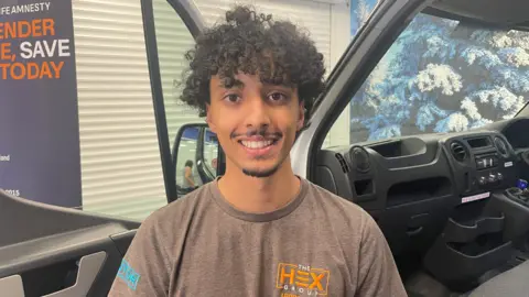 A man with long, curly dark hair and wearing a brown shirt with The Hex Group logo on it, smiles as he stands by the open passenger-side door of a van.