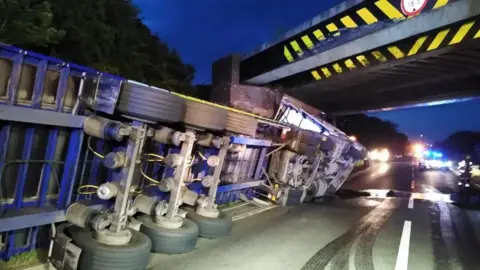 Image shows a lorry overturned on its left side. The front of the lorry is on its side underneath the A5 Watling Street Bridge and the back and you can see the underside of the lorry with its wheels showing. 