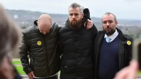 Pacemaker Press Three men stand in a line with their arms around each other at Stormont. They are Declan, Niall and Brendan McNally, brothers of the murder victim. The are each wearing dark jackets. Two of them have beards, the other brother is standing with his head bowed and his face is not fully visible. Two of the brothers are wearing yellow stickers on their jackets. 