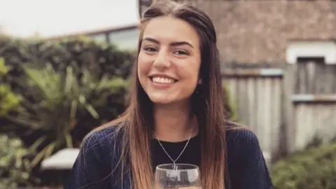 Family photo A young woman with long brown hair smiles at the camera while holding a wine glass. She is wearing a blue long sleeved top with a necklace around her neck. 