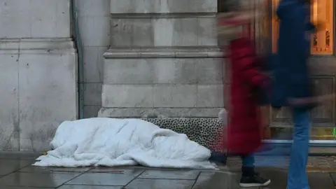 A homeless person underneath a white blanket on a wet street. Two blurred people are walking past