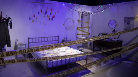 A fictional crime scene with references to films and TV. In the centre there's a bed with lots of air fresheners hanging over it
Behind it is a cordless 90s phone. to the right there's an entrance to a bathroom with a shower curtain drawn
on the wall is a poster of Raquel welch and a tea cup and saucer with a teaspoon in it.
All are movie or tv references
