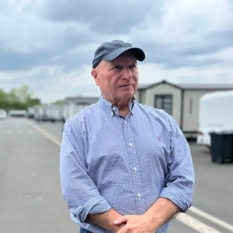 Billy Welch, wearing a blue checked shirt and a baseball cap. He stands with his hands clasped in front of him; a row of caravans can be seen behind him.