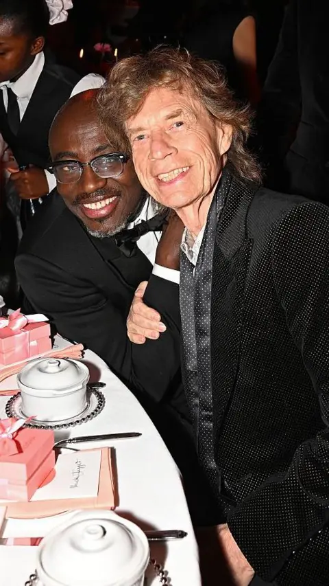 Getty Edward Enninful and Mick Jagger sit together at a table in their suits, smiling