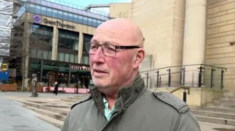 A man with a bald head and glasses is wearing a dark green coat and is standing near the steps of Sheffield City Hall