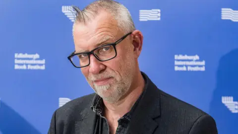 Getty Images A man with thinning, grey, spikey hair and a grey beard and glasses in a dark suit jacket with a black, unbuttoned shirt underneath