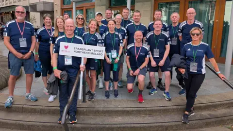 Team NI A team photo of Transplant Sport Northern Ireland. They are all standing on concrete steps. One man near the front is holding a white sign reading: 'Transplant Sport Northern Ireland'
