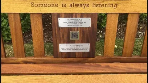 Legend on the Bench A wooden bench. Three small metal plates are inscribed with numbers and a QR code carrying details of mental health support groups.