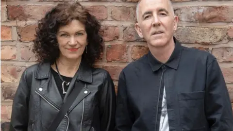 Alison Bell has dark red hair and is wearing a black motor bike jacket. Liam Walsh has greying hair around the temples and is wearing a dark blue jacket