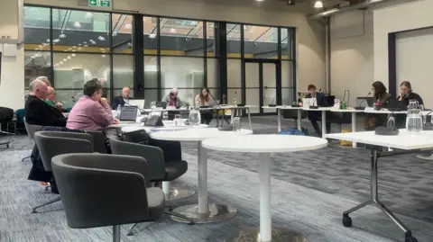 Joe Griffin/LDRS A meeting room Peterborough's Engine Shed. At least 10 women and men can be seen sitting in a horseshoe shape around tables with microphones around them. Behind them is a large window, opening into an interior hall or space in the building. 