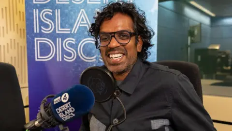 An image of the comedian Romesh Ranganathan with a microphone that has discs from the desert island.