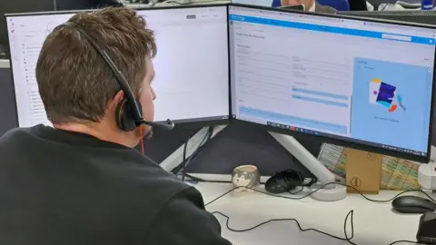 A man sat in front of a computer with a head set. He is wearing a black jumper. 