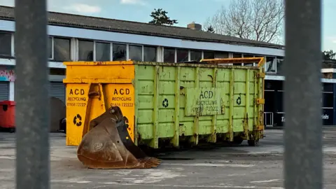 A look through a grey fence, showing a yellow and lime green skip bin with a claw placed to the side 