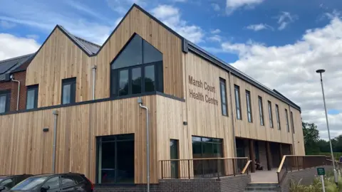 A light brown wood building, with big windows. On the side it reads "Marsh Court Health Centre".