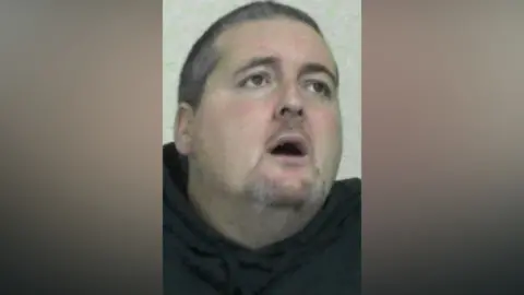 Mugshot of Michael Gill. He has a large head and short dark grey hair. He is looking up and to his left with his mouth wide open and a confused look in his eyes.