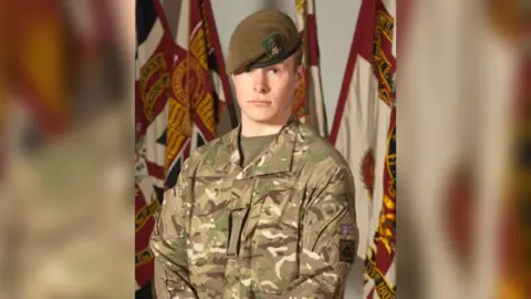 Ministry of Defence Pte Stone, pictured wearing camouflage military fatigues and green beret of The Yorkshire Regiment. The soldier is stern-faced and has the regimental flag behind him.