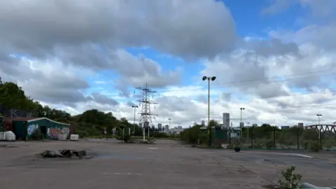 Alexander Brock Land including a building with graffiti on the far left. There is little on the ground in the bottom part of the image, while an electricity pylon is in the background, along with a small part of the Birmingham skyline. 