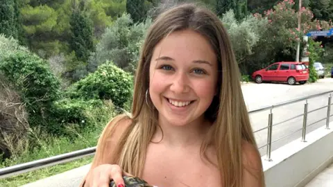 Family Handout Ellie Gould standing on an outdoor balcony with trees and bushes in the background. She has blue eyes, long and straight light brown hair and is wearing silver hoop earrings, smiling at the camera.