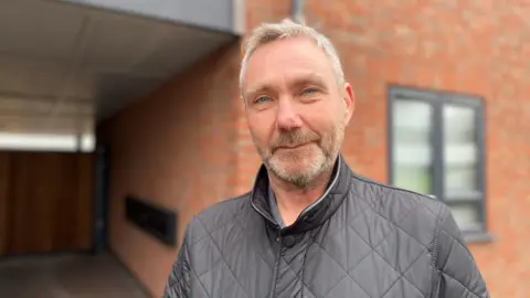 Alan Morris, a man in a black Barber-style coat and white hair/facial hair, standing outside his building