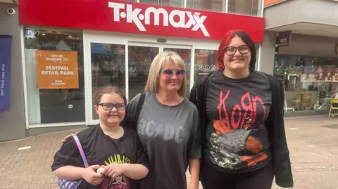 A girl is on the left, a woman with blonde hair is in the middle and a woman with partly red hair is on the right. All three are wearing glasses and standing in front of TK Maxx.