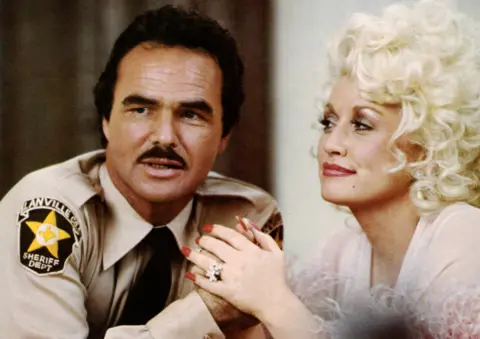 Universal Pictures via Alamy Burt Reynolds in a sheriff's costumes looks straight ahead, while Dolly Parton in with her hair very curly and swept up, looks at him and holds his hands. 