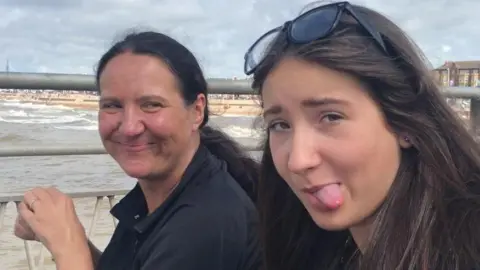 A mum smiles as she looks at her daughter, to her left, pulling a silly face at the camera. They are in the foreground and behind them is the sea and a beach. 