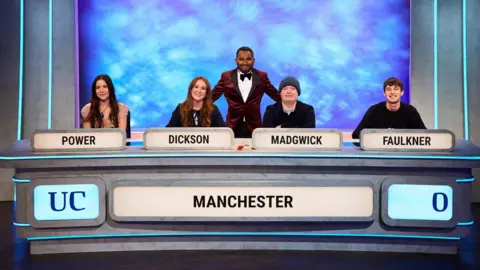BBC/ITV Studios Four people sitting behind a TV quiz show desk, left to right a woman with long dark hair, a woman with long red hair,  a person in a blue beanie, and a man with short dark hair. In front of them are their surnames illuminated - POWER, DICKSON, MADGWICK, FAULKNER and below that MANCHESTER. Between Dickson and Madgwick stands a man in a crimson velvet tuxedo jacket, white shirt and black bow tie, and short black hair. 