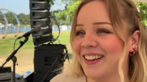 A picture of Emily Wishart on a stage, with a microphone and speakers behind her on a fine summer's day. She has shoulder length blonde hair.