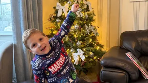 Family photo A young boy wearing a Christmas Marvel jumper and smiling with his hand in the air in front of a Christmas tree