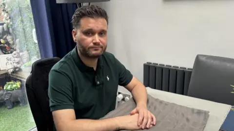 A man with short brown hair and a beard who is wearing a short sleeved green polo shirt looks sad as he sits at a dining table in a house. His hands are placed on what appears to be a pale brown towel placed on top of the table. 