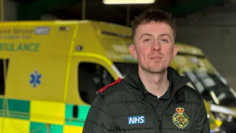 Owen Finney is standing in front of two parked ambulances and smiling slightly at the camera. He has short brown hair, and he is wearing a green uniform with an ambulance crest on it and a blue patch which reads: "NHS".