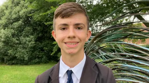 Alex Pope/BBC Charlie smiles directly at the camera, wearing a black blazer, blue tie and white shirt. He is photographed in a garden with green shrubbery behind him. 