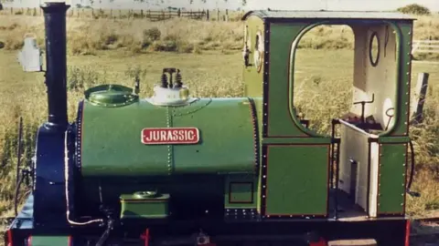 LCLR A sideways view of Jurassic - a green-painted, narrow-gauge, locomotive, with green scrubland in the background 