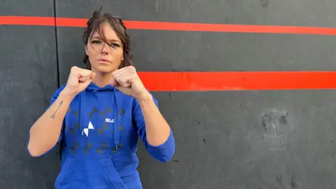 Robyn Wereshcuk who is wearing a blue sports hoodie and is standing against a grey wall with her fists clenched and raised as if ready to fight. She has dark hair that has been blown across her face by the wind.